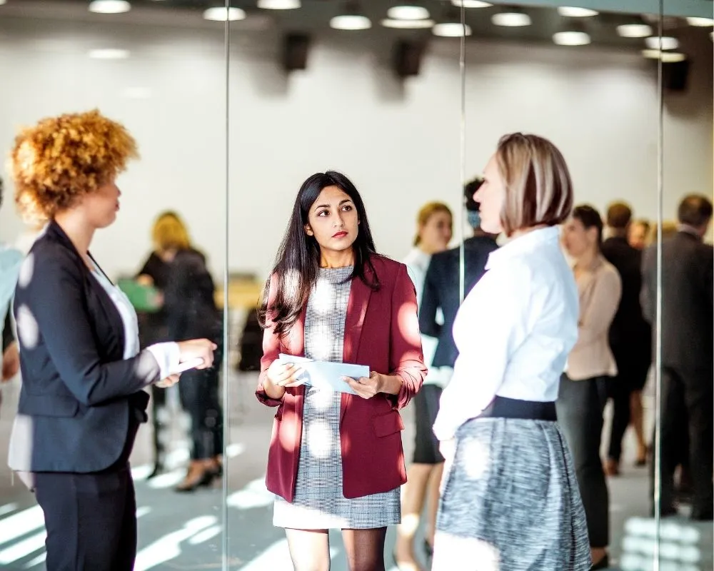Three women in business attire talking at a corporate networking event, with more attendees mingling in a bright conference space behind them