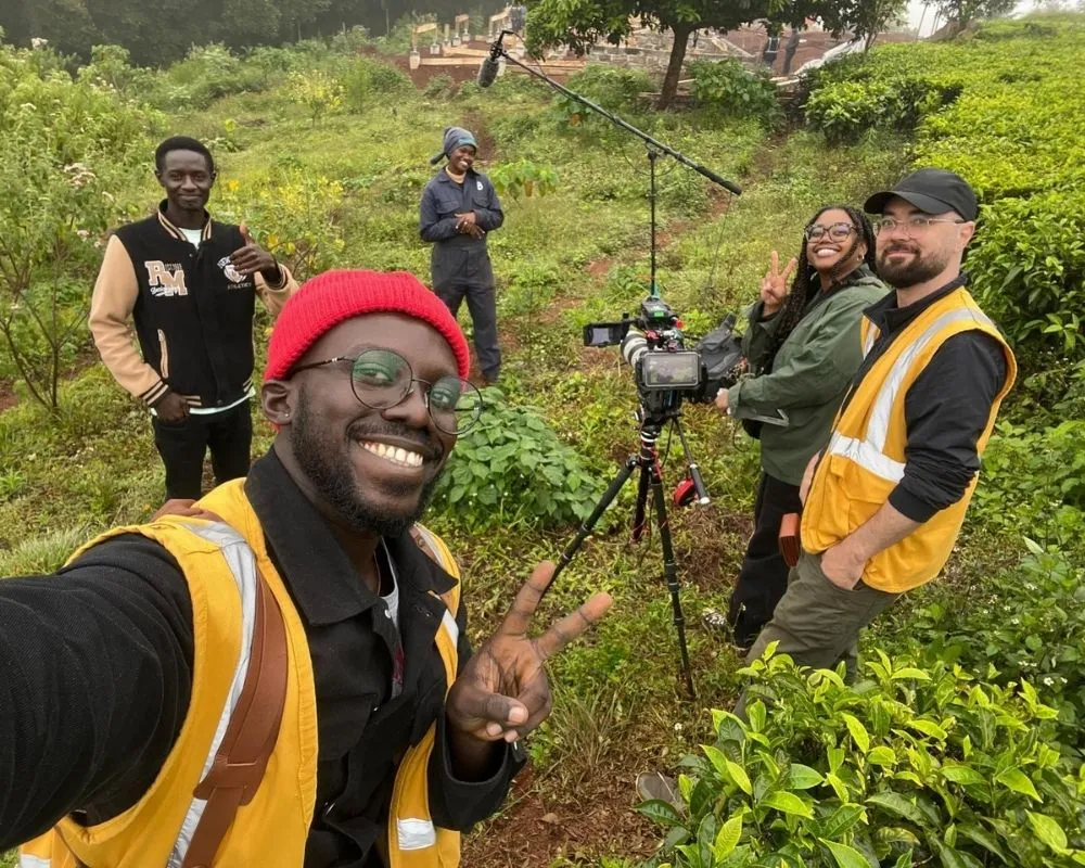 Smiling documentary crew in safety vests on a green hillside, with a tripod camera and boom mic set up to film on location