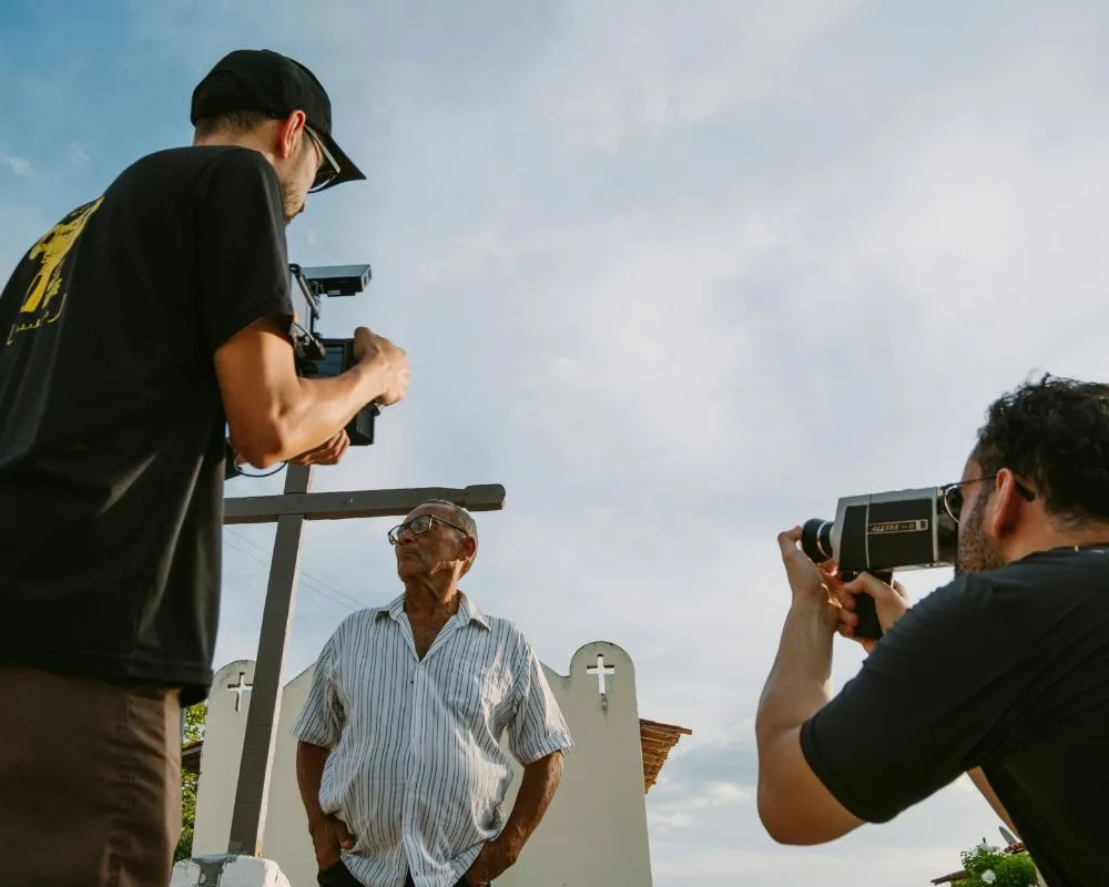 Two camera operators, one holding a vintage film camera, filming an older man beside a wooden cross for a documentary