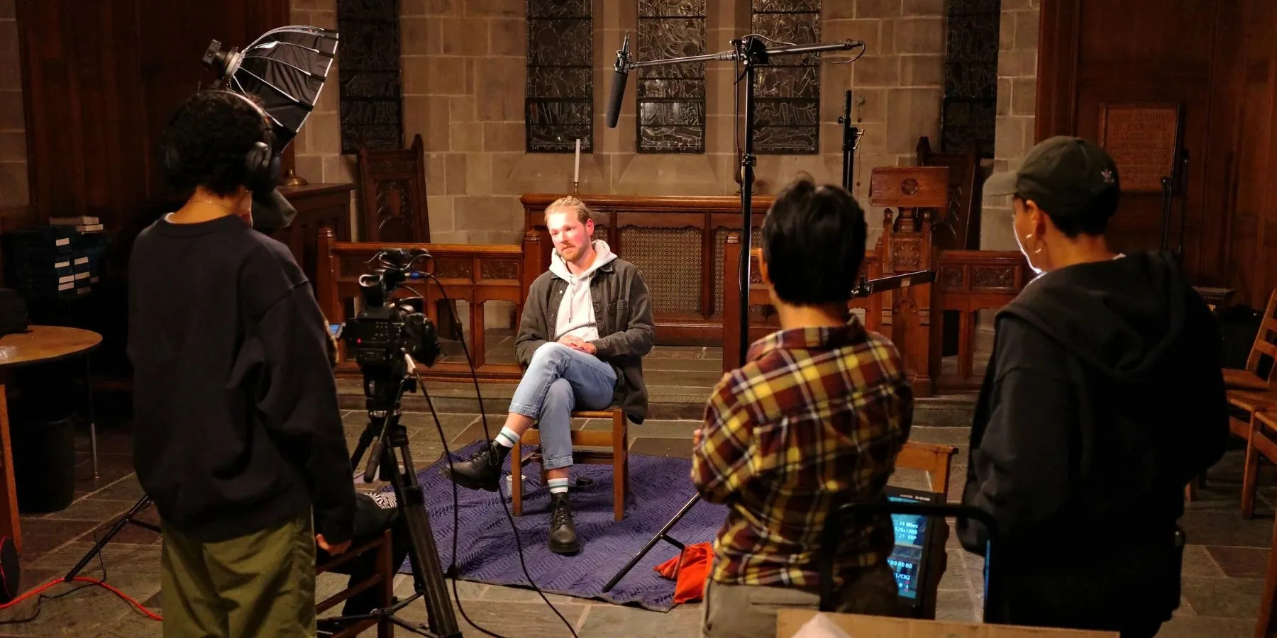 Documentary video production crew filming a seated interview in a stone chapel with a softbox light and boom microphone
