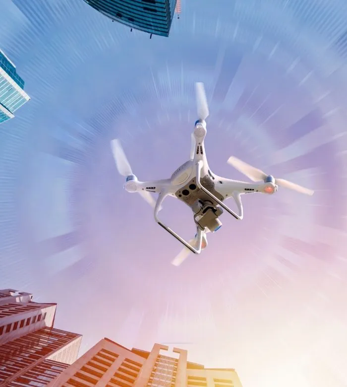 White camera drone flying between tall city buildings, seen from below