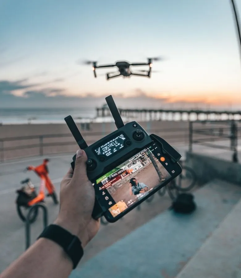 Drone pilot holding a remote controller with a live camera view as a drone flies over the beach at sunset