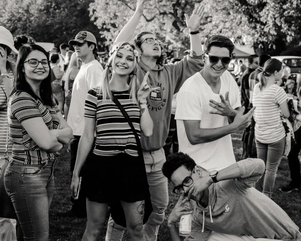 Black and white photo of smiling friends cheering and posing in the crowd at an outdoor festival, one wearing a flower crown