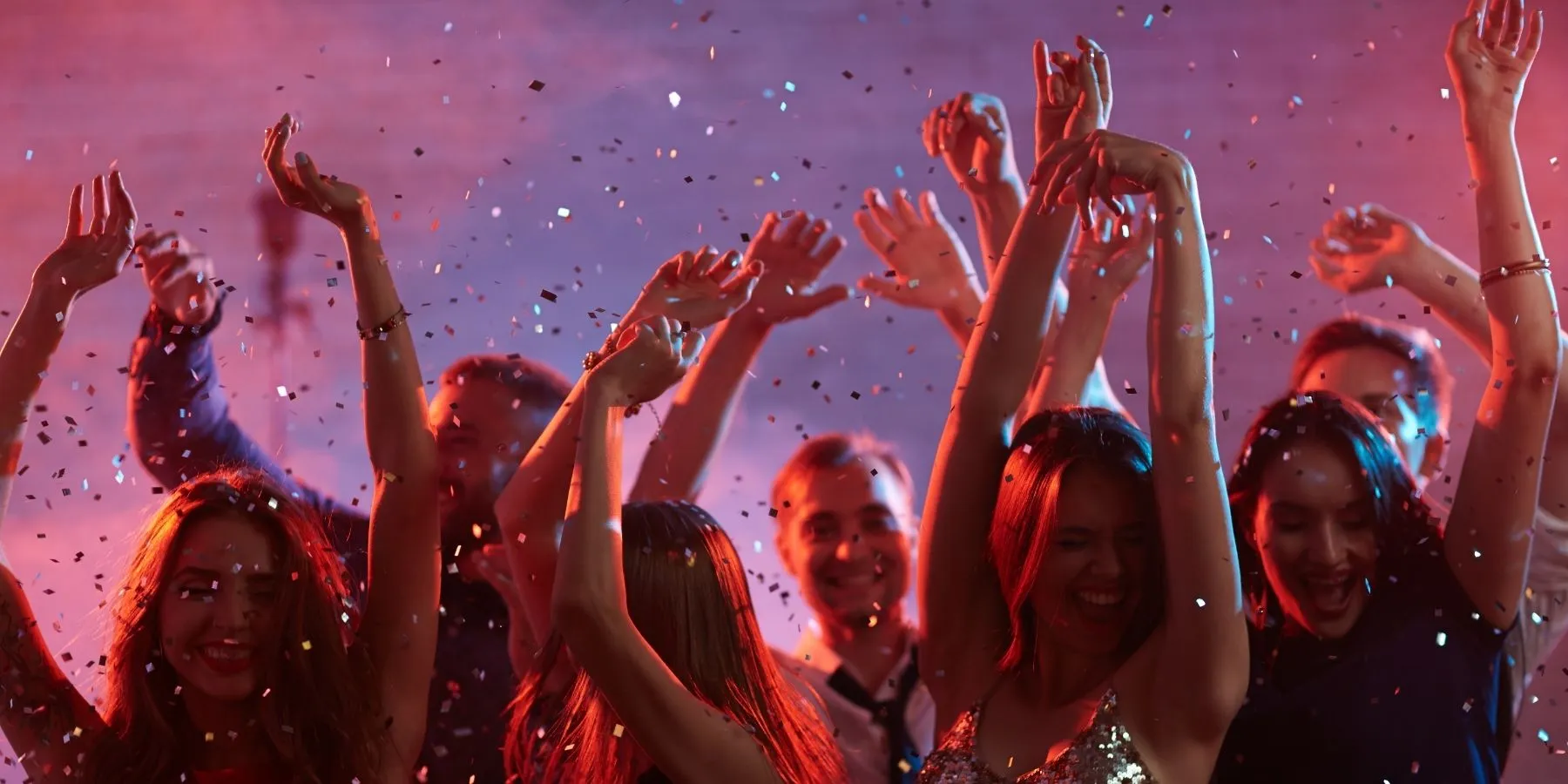 Party guests dancing with raised arms under falling confetti and pink and purple event lighting